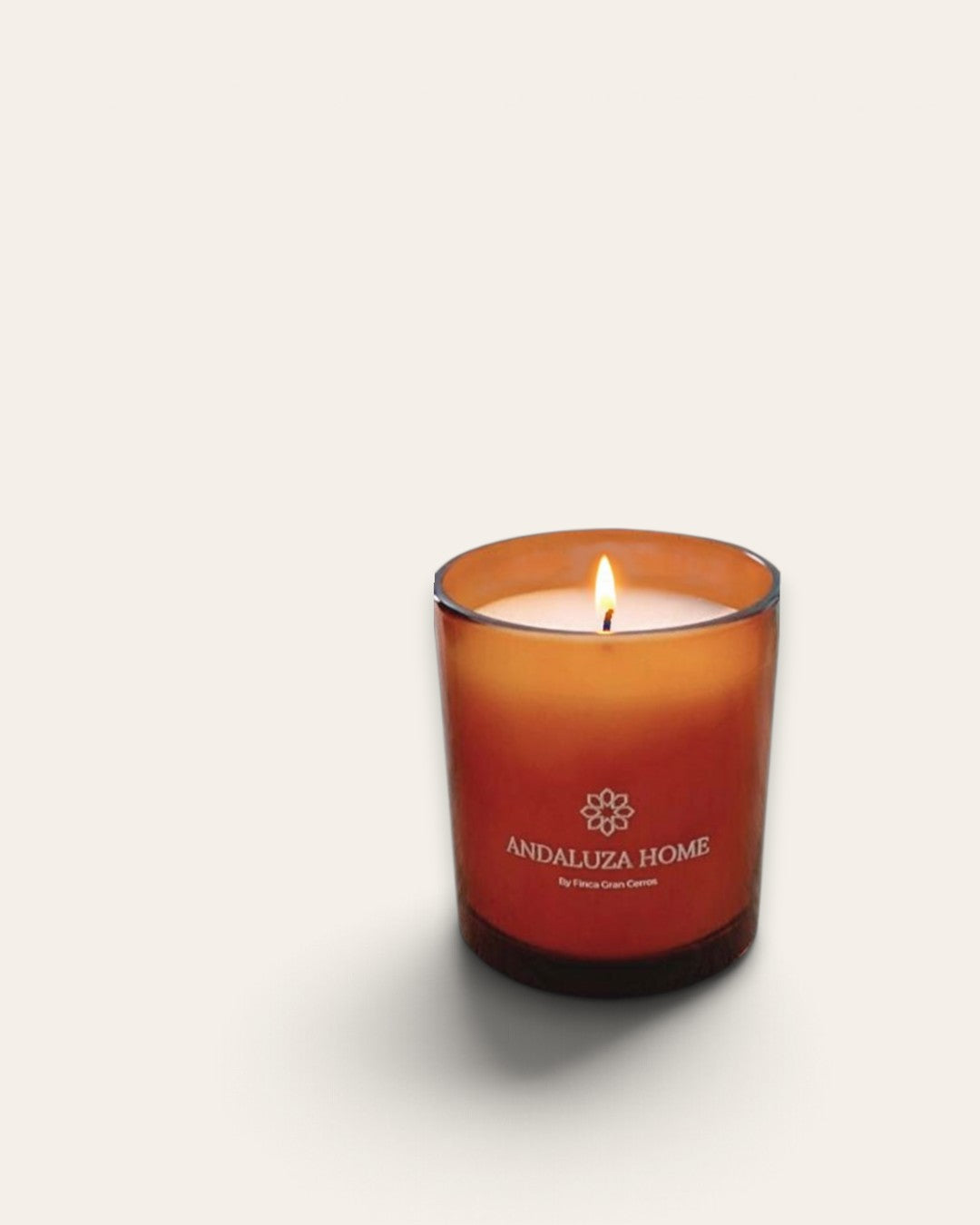 A lit amber glass scented candle with a white wick, featuring the brand name 'ANDALUZA HOME' printed on the front, set against a plain off-white background.
