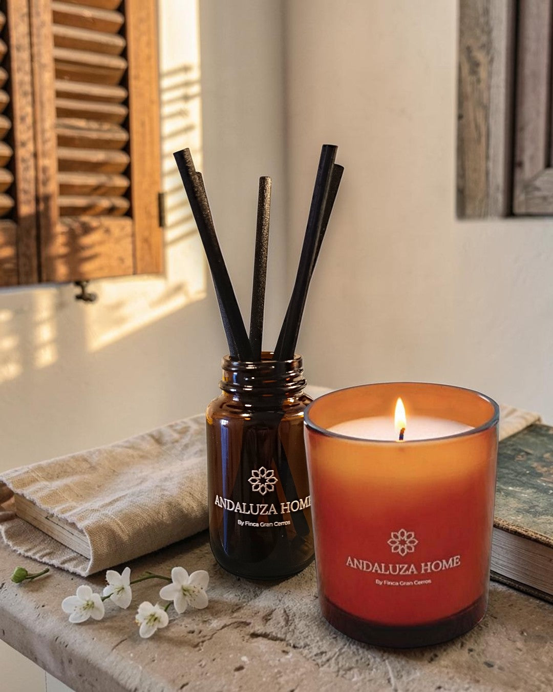 Andaluzia Home reed diffuser and scented candle set on a stone surface with a folded linen cloth, small white flowers, and warm sunlight. The amber glass diffuser jar has black reeds, and the lit orange-red glass candle sits beside it.