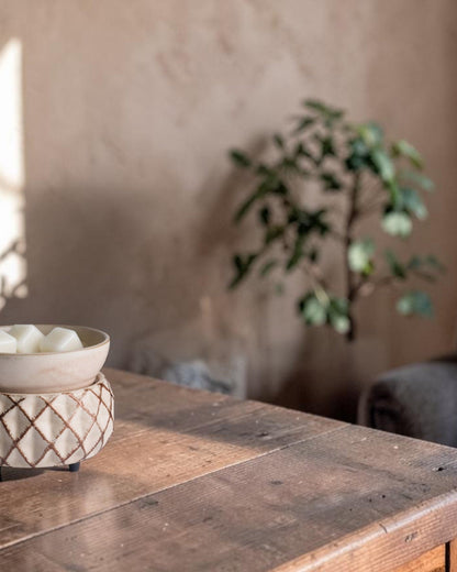 A small beige ceramic bowl with a textured geometric base sits on a rustic wooden table in a softly lit room, with a blurred plant and chair in the background.