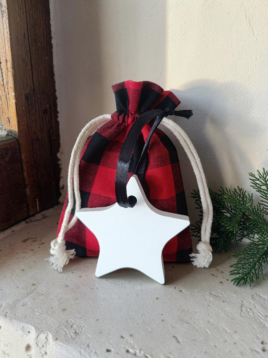A small red and black buffalo plaid drawstring pouch with white rope ties and a black ribbon, holding a white ceramic hanging star ornamental diffuser, styled on a stone ledge with pine branches.